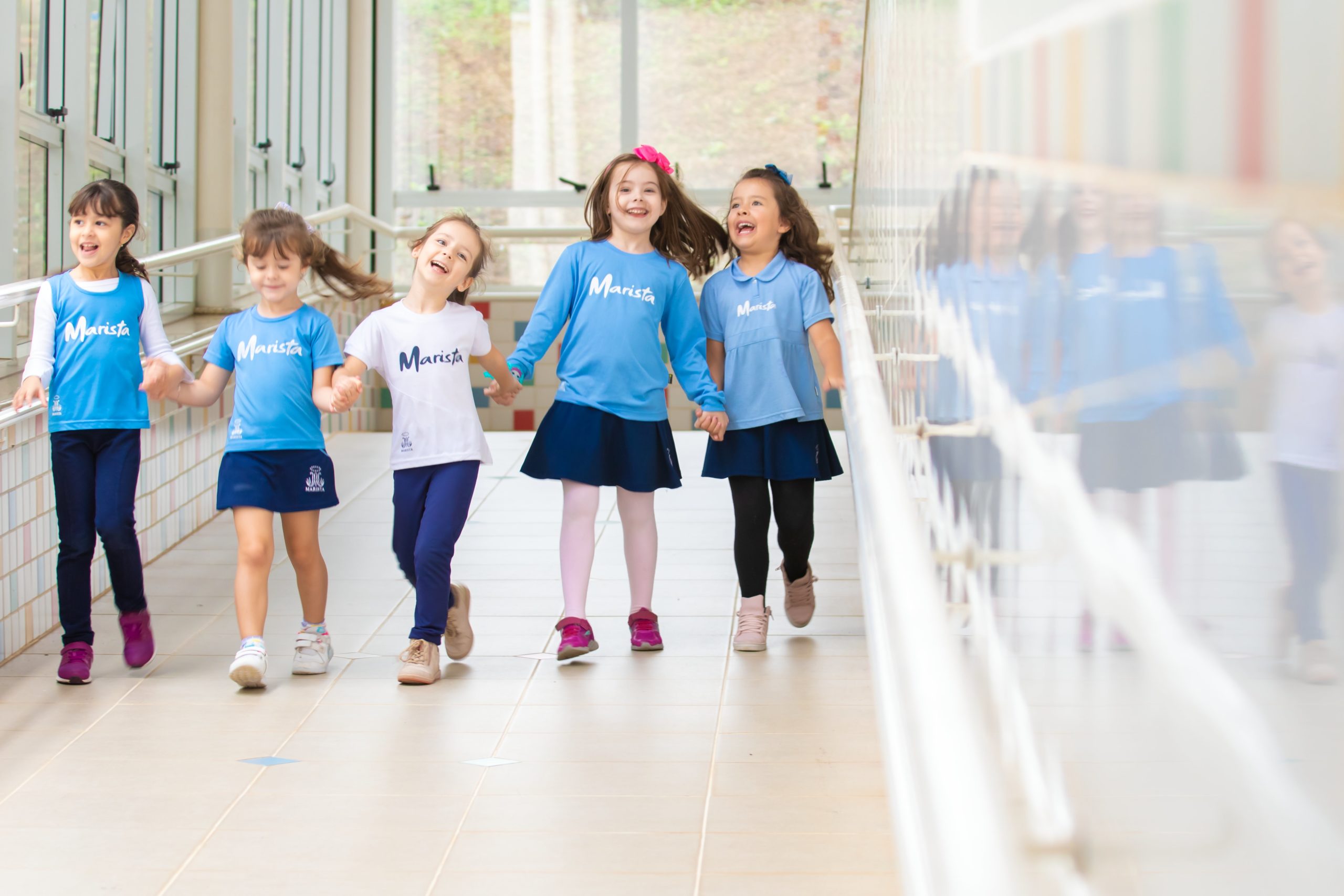 Cinco meninas estudantes com o uniforme do Colégio Marista de mãos dadas em um corredor. 